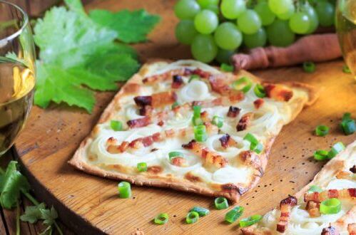 Zwei Gläser Weißwein und Stücke Flammkuchen mit Speck und Zwiebeln auf Holzteller, grüne Weintrauben im Hintergrund. Perfekte Weinbegleitung!