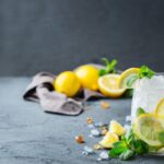 Erfrischender Gin-Cocktail mit Eiswürfeln, Zitronenscheiben und Minzblättern; im Hintergrund liegende Zitronen und ein graues Tuch.