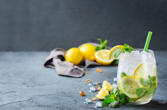 Gin Tonic Glas mit Eis, Zitronenscheiben, Minze und Strohhalm, Zitronen im Hintergrund auf grauem Untergrund. Erfrischung pur!