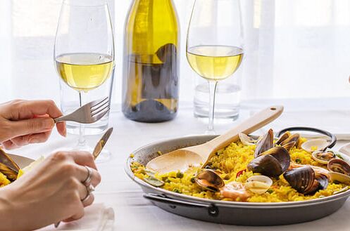 Zwei Personen genießen Paella mit Meeresfrüchten an einem gedeckten Tisch, begleitet von spanischem Weißwein in Gläsern und einer Weinflasche.