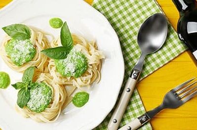 Ein Teller Pasta mit grünem Pesto und eine Flasche Rotwein auf gelbem Tisch mit grünem Tuch. Perfekt für italienisches Abendessen.