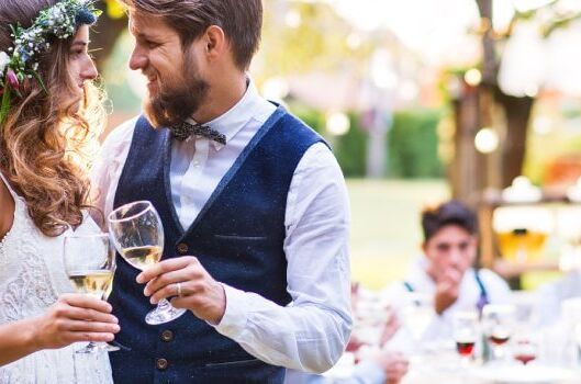 Junges Brautpaar im Freien bei ihrer Hochzeit mit Weingläsern in der Hand, umgeben von feiernden Gästen und festlicher Dekoration. Idealer Hochzeitswein für besondere Momente.