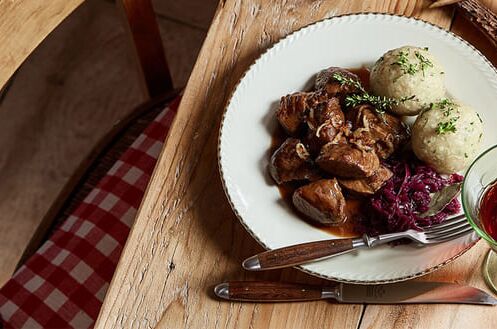 Ein gedeckter Holztisch mit Wildfleisch, Rotkraut und Knödeln auf einem Teller, daneben ein Glas Rotwein