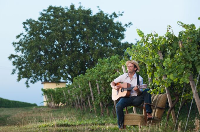 Junger Mann auf Sessel, spielt Gitarre inmitten der Reben des Bioweinguts Lorenz in Deutschland. Genießen Sie Natur und Musik!