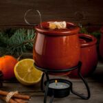 Orangene Feuerzangenbowle-Töpfe und Zutaten wie Orangen, Zimtstangen und Teelicht auf Holztisch. Perfekt für Weihnachtspartys.