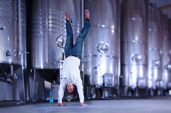 Ein Winzer vom Bioweingut Lorenz steht auf den Händen vor großen Edelstahltanks in einer Weinkellerei, neben einer Flasche und einem Glas Weißwein.