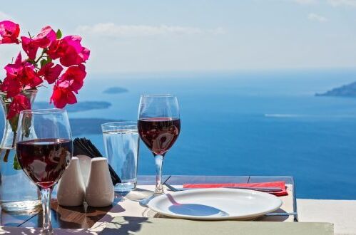 Zwei Gläser griechischer Rotwein auf einem Tisch mit Vase und Blumen, mit Blick auf das blaue Meer und die Inseln im Hintergrund.