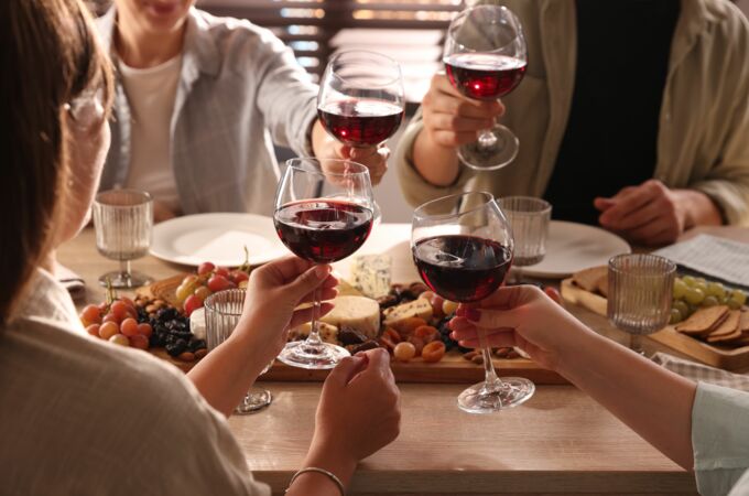 Eine Gruppe von Menschen stößt mit Gläsern voll rotem Bordeaux-Wein an, während sie an einem gedeckten Tisch sitzen. Auf dem Tisch befinden sich verschiedene Käsesorten, Trauben, Nüsse und Trockenfrüchte.