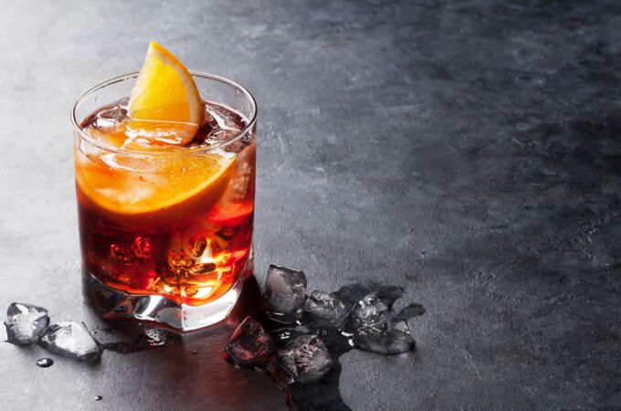 Ein Glas Negroni-Cocktail mit Gin, rotem Vermouth und Campari, garniert mit einer Orangenscheibe auf einer Steinoberfläche mit Eiswürfeln. Perfekter Drink für Genießer!