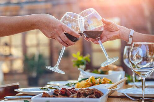 Zwei Personen stoßen mit Rotweingläsern an, im Hintergrund ein reich gedeckter Tisch im Freien bei Sonnenuntergang. Genießen Sie Ihren Saint-Emilion Rotwein!