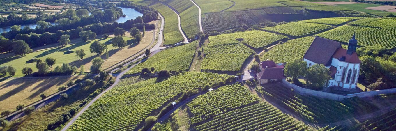 Luftaufnahme des Weingut Zur Schwane in Deutschland, umgeben von ausgedehnten Weinbergen und einer Kapelle unter klarem blauen Himmel.