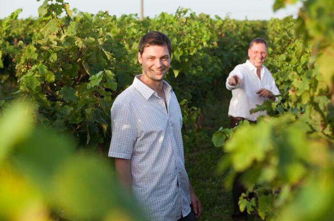 Zwei Winzer, einer im Vordergrund mit hellem Hemd lächelnd, der andere im Hintergrund in einem Weinberg des Bioweinguts Lorenz.