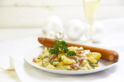 Traditionelles Weihnachtsessen mit Kartoffelsalat und Wiener Würstchen, dekoriert mit einem Glas Weißwein im Hintergrund.