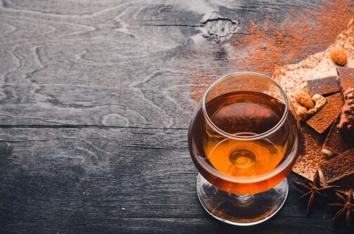 Ein Glas Cognac Weinbrand auf einem dunklen Holztisch mit Schokoladenstücken, Nüssen und Gewürzen im Hintergrund. Genieße den feinen Genuss von Spitzenqualität!