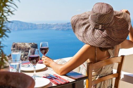 Frau mit Sonnenhut genießt Agiorgitiko Rotwein auf einer Terrasse mit Blick auf das Meer in Griechenland