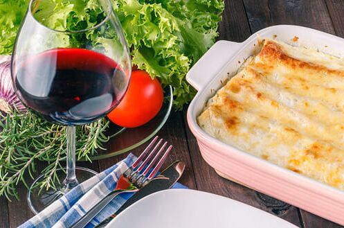 Ein Glas Rotwein neben einer Auflaufform mit Lasagne, frischem Gemüse und Kräutern auf Holztisch. Perfekt für ein köstliches Abendessen.