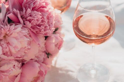 Roséwein im Glas neben rosa Pfingstrosenblüten, sommerlich arrangiert auf weißem Stoff.