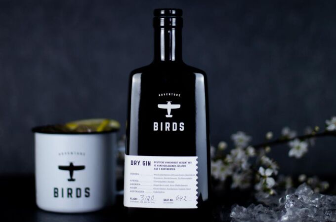 Schwarze Flasche Craft Circus Birds Dry Gin mit weißem Becher und Zitrone, neben Eis und blühenden Zweigen, auf dunklem Hintergrund. Ideal für Gin-Liebhaber.