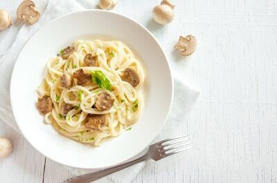 Ein Teller mit cremiger Pasta, garniert mit frischen Champignons, auf weißem Hintergrund. Perfekter Wein-Begleiter für Pilzgerichte.