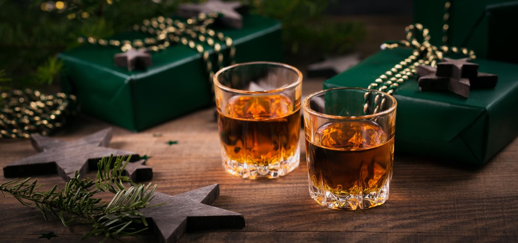 Zwei Whiskygläser auf Holztisch mit grünen Weihnachtsgeschenken und Dekoration, perfekt für festliche Anlässe.