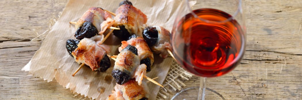 Ein Glas roter Portwein neben Tapas aus Speck und Dörrpflaumen auf rustikalem Holzbrett. Genuss für besondere Anlässe!