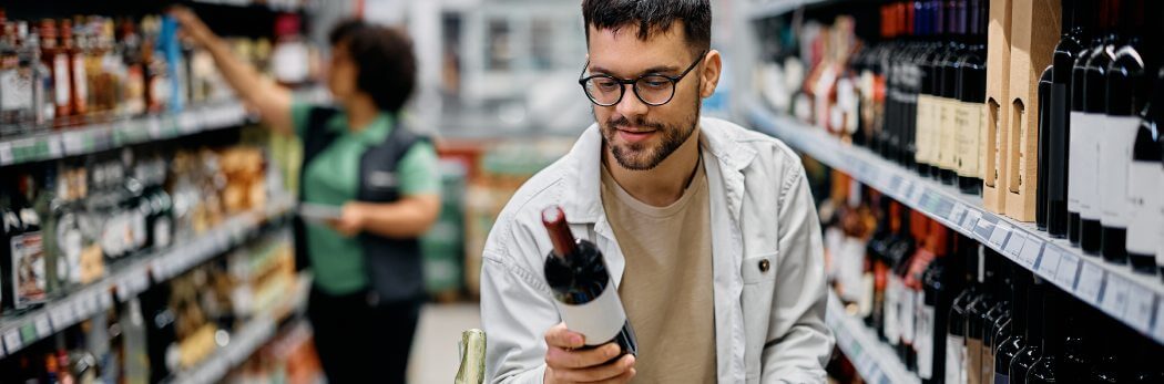 Ein junger Mann mit Brille und Bart betrachtet eine Flasche Rotwein im Weinregal eines gut sortierten Geschäfts. Ideal für Weinanfänger.