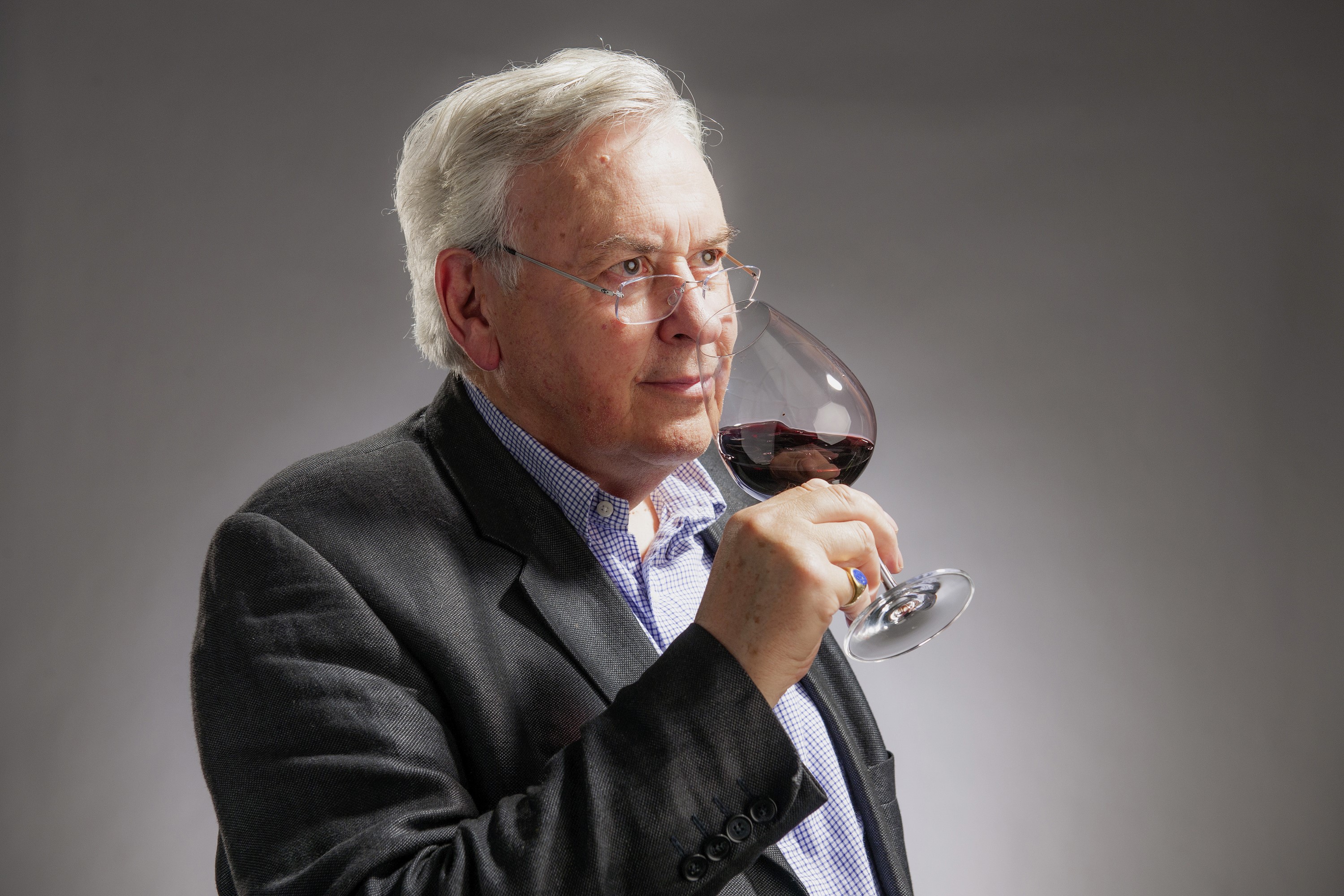 Älterer Mann mit grauen Haaren und Brille kostet in einem Weinglas Rotwein. Auf grauem Hintergrund. Genussvoller Moment im Weinkeller.